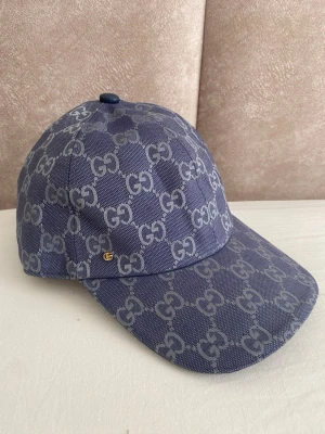 Blå keps från Gucci med GG-mönster - Snygg blå keps från Gucci med det klassiska GG-monogrammet i ljusgrått över hela kepsen. Kepsen har böjd skärm och justerbar passform. Tillverkad i ett slitstarkt tyg med insida i beige bomull. Perfekt för dig som vill ha en exklusiv och trendig accessoar.