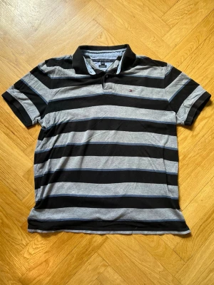 Randig pikétröja från Tommy Hilfiger - Snygg pikétröja från Tommy Hilfiger med breda svarta och gråa ränder. Klassisk krage och korta ärmar, tillverkad i mjuk bomull. Liten logga på bröstet och normal passform. Perfekt för en avslappnad och stilren look.