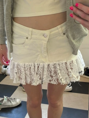 Vit kortkjol med spetsvolang - Supersöt vit kortkjol med femficksmodell och bred volang i spets längst ner. Kjolen har en klassisk jeanslook upptill och spetsdetaljen ger en romantisk vibe. Perfekt för dig som vill sticka ut lite extra. Har lagt till en extra knapp!💕💕💕