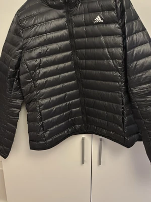 Svart dunjacka från Adidas - Snygg svart dunjacka från Adidas med huva och dragkedja framtill. Jackan har ett diskret Adidas-logga på bröstet och quiltad design med horisontella sömmar. Perfekt för kalla vinterdagar och ger en sportig look.
