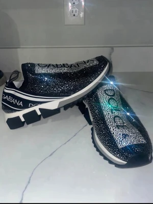 Dolce & Gabbana glittriga sneakers - Säljer ett par svarta Dolce & Gabbana sneakers täckta med glittrande strass och tydliga logotexter på ovansidan. Skorna har chunky vit och svart sula, elastisk slip-on design och sportig look. Perfekta för dig som vill sticka ut med exklusiv stil.