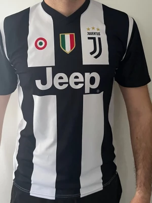 Juventus matchtröja Ronaldo #7 - Juventus fotbollströja i svart och vitt med klassiska ränder, Jeep-logga på bröstet och Ronaldo #7 på ryggen. Tröjan har klubbmärke, italienska flaggan och två stjärnor ovanför loggan. Korta ärmar och sportig passform, perfekt för fotbollsträning eller support. 