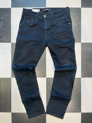 Replay Anbass W31 L33 - Snygga mörkblå slim fit jeans från Replay med klassisk femficksmodell och diskreta slitningar framtill. Jeansen har normal midja, smala ben och är tillverkade i stretchigt denim för extra komfort. Perfekta för en trendig och avslappnad look.