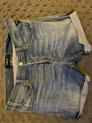 Blå jeansshorts från Replay - Säljer ett par blå jeansshorts från Replay med uppvikta benslut och klassisk femficksmodell. Shortsen har Hyperflex-material som ger extra stretch och komfort. Perfekta för sommaren och har snygga slitningar för en avslappnad look.