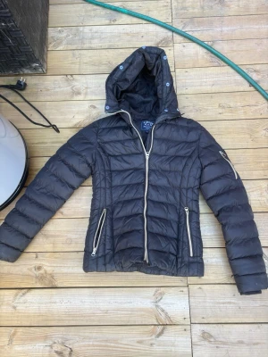 Svart dunjacka från Vinson Polo Club - Snygg svart dunjacka från Vinson Polo Club med huva och dragkedjor i kontrasterande beige. Jackan har två fickor framtill med dragkedja och en extra ficka på ärmen. Perfekt för kalla vinterdagar och har en modern, figurnära passform.