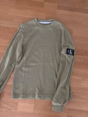 Olivgrön långärmad tröja Calvin Klein - Olivgrön långärmad tröja från Calvin Klein med diskret struktur i tyget och CK-logga på ärmen. Tröjan har rund halsringning och en relaxed passform, perfekt för en avslappnad stil.
