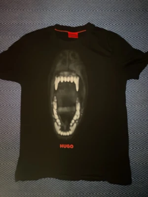 Svart t-shirt med tryck från HUGO - Cool svart t-shirt från HUGO med ett stort tryck av ett öppet djurgap på bröstet och röd HUGO-logga under. Klassisk rund hals och korta ärmar. Perfekt för dig som vill sticka ut med en edgy look.