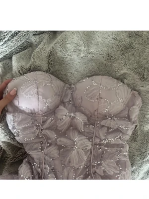 Ljus lila axelbandslös klänning - Axelbandslös kortklänning i ljus lila med blommigt mönster och paljetter. Klänningen har en tight passning och är dekorerad med broderade blommor och glittrande detaljer. Perfekt för dig som vill sticka ut med en unik look.