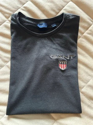 Svart t-shirt från GANT med logga - Svart t-shirt från GANT med broderad logga och emblem på bröstet. Klassisk rund halsringning och korta ärmar. Material bomull 