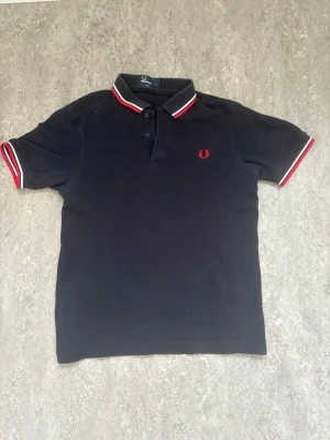 Svart Fred Perry pikétröja - Säljer en svart pikétröja från Fred Perry med klassisk krage och knappar. Tröjan har röda och vita detaljer på krage och ärmslut samt Fred Perry-loggan broderad i rött på bröstet. Perfekt för dig som gillar stilrena och sportiga plagg.