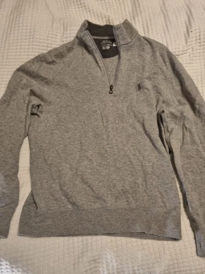 Grå halvzip från Polo Ralph Lauren - Snygg grå tröja med halv dragkedja från Polo Ralph Lauren. Tröjan har broderad logga på bröstet, hög krage och mjukt material. Perfekt för lager-på-lager och enkel att matcha med jeans eller chinos.