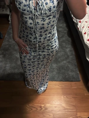 Vit långklänning med blå blommor - Supersöt långklänning i vit med blått blommönster. Klänningen har smala axelband med knyt, v-ringning och figurnära passform. Perfekt för sommarens alla tillfällen och ger en romantisk vibe. Materialet känns mjukt och svalt, troligen bomull eller polyester.