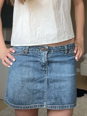 Blå jeanskjol från Wrangler - Säljer en klassisk blå jeanskjol från Wrangler i modellen Kylie. Kjolen är i 100% bomull, har normal passform, bälteshällor och knappstängning framtill. Perfekt till sommaren och enkel att styla med olika toppar.