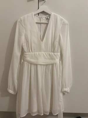 Vit omlottklänning med v-ringning - Snygg vit klänning med lång ärm, markerad midja och v-ringad hals. Klänningen har omlottkänsla i midjan och är tillverkad i ett lätt, flowy material som ger en luftig känsla. Perfekt för sommarens tillfällen.