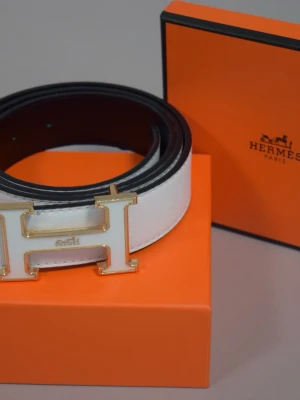 Vitt bälte med H-spänne - 120cm. Stilrent vitt bälte med ikoniskt H-spänne i guld och vitt. Skärpet är gjort i exklusivt skinn och har en klassisk, tidlös design som lyfter vilken outfit som helst. Perfekt accessoar för dig som gillar lyxiga detaljer.