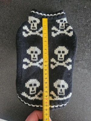 Stickad hundtröja med dödskallar - Svart stickad hundtröja med vita dödskalle- och korsade ben-motiv. Tröjan har hål för frambenen och ribbade kanter vid hals och ben. Mjuk och stretchig, perfekt för att hålla din hund varm och snygg.