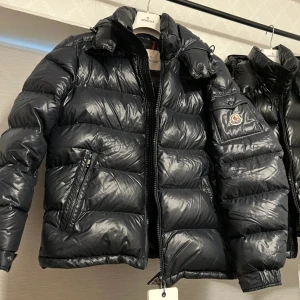 Svart Moncler Maya jacka  - Säljer en svart pufferjacka från Moncler med glansig finish och klassisk logga på ärmen. Jackan har hög krage, två sidofickor med dragkedja och quiltad design. Perfekt för dig som vill ha en snygg och varm jacka med exklusiv känsla. Bästa jackan att köpa inför vintern.