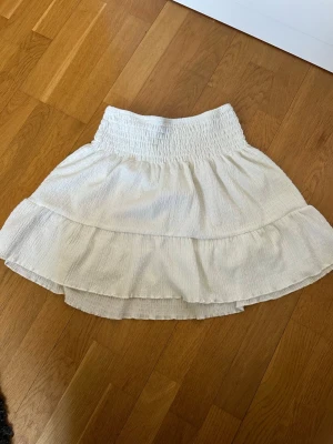 Vit volangkjol från Gina Tricot - Supersöt vit kortkjol från Gina Tricot med bred smock i midjan och dubbla volanger. Kjolen har en luftig och lätt struktur, perfekt för varma dagar. Enkel att matcha med både toppar och sneakers för en fräsch look.