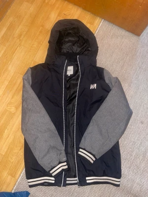 Svart och grå bomberjacka med huva - Cool bomberjacka från Warp med svart kropp och grå ärmar. Jackan har huva. Materialet är troligtvis polyester och bomull, perfekt för att ge en sportig look.