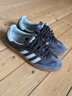 Bruna Adidas Samba - Klassiska Adidas Samba sneakers av brun läder med vita ränder och gråa beiga mockadetaljer. Gummisula och brun snörning. Ikonisk retrostil med guldfärgad SAMBA-text på sidan. Har haft de i cirka en månad