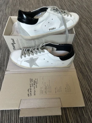 Vita golden goose - Säljer ett par vita Golden Goose sneakers med grå mockastjärna på sidan, grå skosnören och svart detalj på hälen. Skorna har en klassisk låg siluett och är tillverkade i läder med mockadetaljer. Perfekta för dig som gillar en avslappnad men trendig stil.