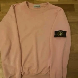 Ljusrosa sweatshirt från Stone Island - Säljer en ljusrosa sweatshirt från Stone Island med den klassiska svarta logotypen på vänster ärm. Tröjan har rund halsringning, ribbade muddar och är tillverkad i mjukt bomullsmaterial. Perfekt för en avslappnad streetwear-look.