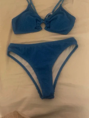 Blå bikini med ringdetalj - Säljer en blå bikini med struktur i tyget och en snygg ringdetalj framtill på toppen. Bikinin har dubbla axelband och hög midja på underdelen. Perfekt för strandhäng eller poolparty!