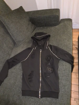 Svart hoodie med döskallepatch - Cool svart hoodie från Philipp Plein med silverfärgad dragkedja och flera unika patchar, bland annat en hexagonformad döskalle. Huva med detaljerade sömmar och ribbade muddar. Perfekt för dig som gillar streetstyle och edgy design. Använd max 3 gånger då en kedja gick sönder i nacken, och kedjan går att fixa om nån orkar. Annars så gott som ny. Kvitto finns, köpt på KOW.