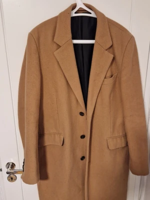 Asos Kappa man medium - Stilren kamelfärgad ullkappa med tre knappar framtill, breda slag och två fickor med lock. Kappan har en rak passform och svart innerfoder, perfekt för en trendig och tidlös look. Passar lika bra till jeans som till kostym.