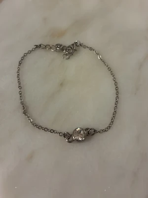 Silverfärgat armband med kristall - Ett stilrent armband i silverfärgad metall med en tunn kedja och en rund, gnistrande kristall i mitten. Armbandet har justerbar längd med flera öglor och ett klassiskt lås. Perfekt för att ge en elegant touch till din outfit.