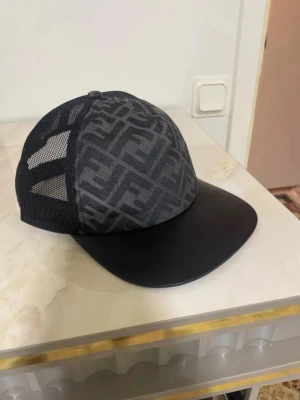 Svart keps med mesh och Fendi-mönster - Snygg svart keps från Fendi med ikoniskt mönster på framsidan och mesh på sidorna för extra ventilation. Böjd skärm i svart och justerbar passform bak. Perfekt för dig som vill ha en stilren accessoar med lyxig känsla.