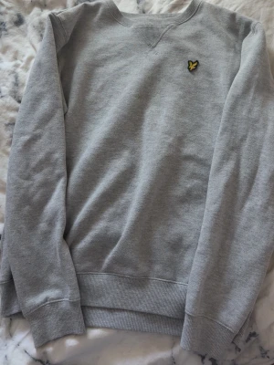 Grå sweatshirt från Lyle & Scott - En stilren grå sweatshirt från Lyle & Scott med deras klassiska gula logga broderad på bröstet. Tröjan har rund halsringning, ribbade muddar vid ärmslut och nederkant samt en relaxed passform. Perfekt för en casual och avslappnad look.