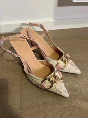 Beige och rosa pumps med gulddetaljer - Säljer ett par eleganta pumps i beige tyg med ljusrosa detaljer och spetsig tå. Skorna har justerbar rem runt hälen och en dekorativ guldfärgad kedja framtill. Klacken är medelhög och bred för extra stabilitet. Perfekta för dig som gillar en chic och trendig look.