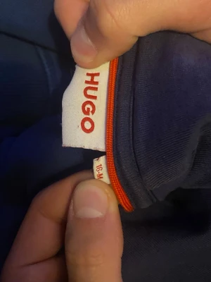 Mörkblå ziptröja från HUGO BOSS - Mörkblå ziptröja från HUGO BOSS med hel dragkedja och långa ärmar. Tröjan har ribbade muddar och en diskret orange detalj vid dragkedjan. Perfekt för dig som gillar stilrena och moderna plagg.