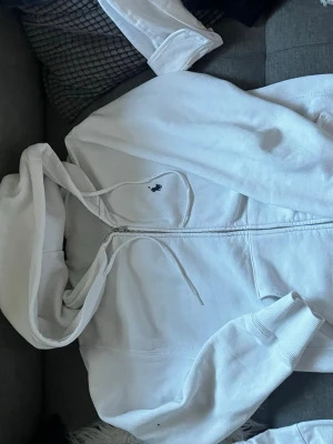 Vit hoodie från Polo Ralph Lauren - Klassisk vit hoodie från Polo Ralph Lauren med dragkedja framtill och justerbar huva. Tröjan har ribbade muddar vid ärmar och nederkant samt den ikoniska lilla broderade loggan på bröstet. Tillverkad i mjukt bomullsmaterial för extra komfort.