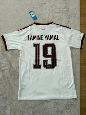 Spanien Lamine Yamal matchtröja - Vit Spanien fotbollströja från Adidas med Lamine Yamal och nummer 19 på ryggen. Tröjan har vinröda detaljer, klassiska Adidas-ränder på axlarna och landslagets emblem på bröstet. Tillverkad i lätt och ventilerande material, perfekt för fotbollsträning eller match.