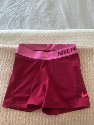 Nike Pro shorts - Snygga och sportiga Nike Pro shorts i en mörkrosa färg med ljusrosa resår i midjan där det står 'NIKE PRO'. Tight passform och tillverkade i stretchigt syntetmaterial, perfekta för träning eller gymmet.