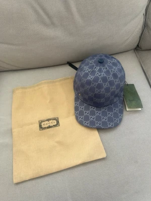 Blå GG-monogram keps från Gucci - Säljer en blå keps från Gucci med det klassiska GG-monogrammönstret över hela kepsen. Kepsen har böjd skärm och justerbar rem bak. Materialet är canvasliknande och kepsen kommer med original dustbag och tagg kvar.