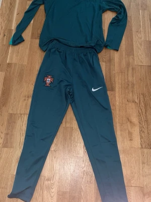 Portugal Nike träningsset - Säljer ett mörkgrönt träningsset från Nike med Portugal-emblem. Setet består av långärmad tröja med halv dragkedja och matchande träningsbyxor. Båda delar har Nike-logga och Portugal-märke. Materialet är lätt och andas, perfekt för fotbollsträning.