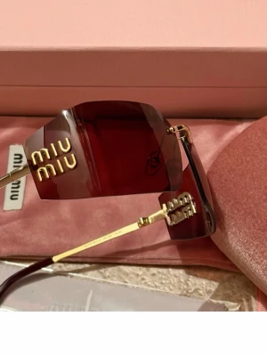 Bruna solglasögon från Miu Miu - Snygga solglasögon från Miu Miu med röda, rektangulära glas och guldfärgade detaljer. Glasögonen har en modern, kantig design och logotypen i guld på sidan av glaset. Perfekt accessoar för dig som vill sticka ut.
