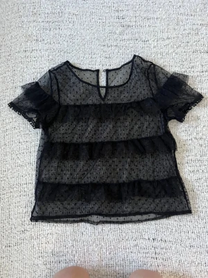 Svart transparent volangblus - Snygg svart blus i transparent mesh med prickigt mönster och volangdetaljer. Korta ärmar med dekorativ kant och liten knapp i nacken. Perfekt att styla över ett svart linne eller en bralette! Storleken är xxs men passar bra också på mig som är xs!🥰