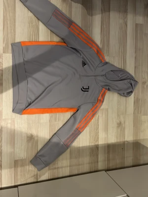 Grå Juventus hoodie från Adidas - Snygg grå hoodie från Adidas med Juventus-logga på bröstet och orangea detaljer längs ärmarna. Klassisk huva och dragkedja vid halsen. Perfekt för dig som gillar sportig stil och vill sticka ut med färgade ränder.
