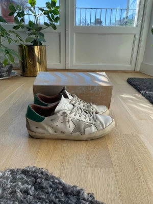 Golden Goose vita sneakers med grön häl - Säljer ett par vita Golden Goose sneakers med ikonisk stjärndetalj på sidan och grön häl. Skorna har låga skaft, vita snören och en vintage-inspirerad look. Tillverkade i läder med slitna detaljer för en cool och avslappnad stil. Rätt så använda därför låga priset 