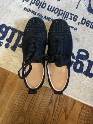 Svarta Louboutin sneakers med nitar - Säljer ett par svarta sneakers från Christian Louboutin med coola svarta nitar på tån och klassisk röd sula. Skorna har snörning och är tillverkade i mocka och läder. Perfekta för dig som vill sticka ut med en lyxig och edgy stil. En spik är borta men annars är den bra lite skitiga men går och tvätta lätt. Använd max 5 gånger 