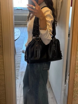 Vintage handväska - Säljer min super fina svarta handväska i skinnimitation med breda axelremmar och snygga metalldetaljer. Köpte i en vintagebutik i Italien. Väskan har två dragkedjor framtill och stora ringar i mässing. Rymlig storlek, får plats med mycket i💞