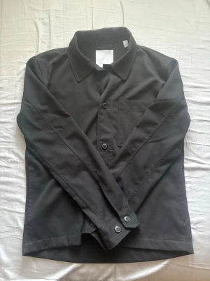 Only&Sons Svart overshirt  - Stilren svart overshirt från Only&Sons, med klassisk krage och knäppning framtill. Jackan har en bröstficka på vänster sida och långa ärmar med knappar vid ärmslut.