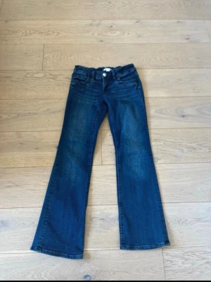 Mörkblå bootcut jeans - Säljer ett par mörkblå bootcut jeans med klassisk femficksdesign och knappgylf. Jeansen har lätt utsvängda ben och är tillverkade i ett mjukt denimtyg. 