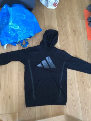 Svart hoodie från Adidas - Svart hoodie från Adidas med stor logga på bröstet i grått. Tröjan har lång ärm, huva och en stor magficka framtill. Materialet är mjukt och passar perfekt för sport eller chill. Snygga detaljer längs sidorna ger ett stilrent intryck.