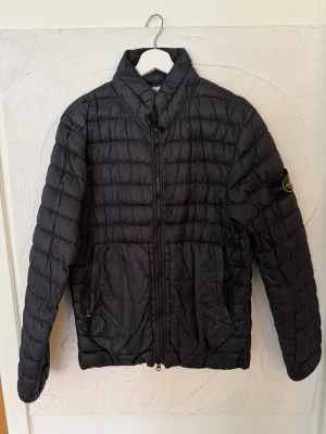Svart pufferjacka från Stone Island - Snygg svart pufferjacka från Stone Island med klassisk patch på ärmen. Jackan har quiltad design, hög krage och dragkedja framtill. Perfekt för kyliga dagar och ger en stilren look. Tillverkad i lätt och smidig polyester.