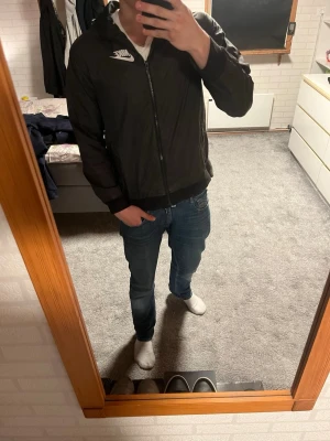 Svart Nike vindjacka med huva - Snygg svart vindjacka från Nike med vit logga på bröstet. Perfekt för blåsiga dagar och enkel att matcha med jeans eller träningskläder. Modellen är 175 cm. Skriv vid frågor, jag svarar snabbt!